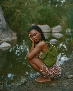 a woman sitting on the ground next to a body of water with her hand under her face