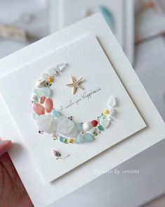 a hand holding up a card with flowers and shells in the shape of a wreath