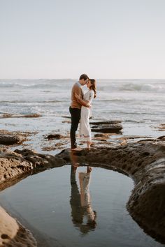 a man and woman standing on rocks near the ocean with their arms around each other