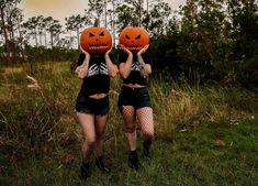 two women wearing halloween costumes holding pumpkins over their heads in a field with trees and grass behind them