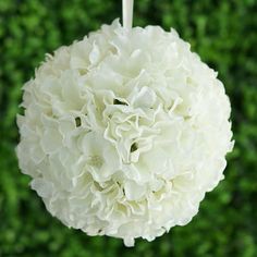 a white flower hanging from a string in front of a green wall with bushes behind it