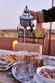 a person pours tea from a kettle into glasses on a table with food and drinks