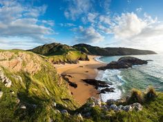 the beach is surrounded by green hills and blue water with white clouds in the sky