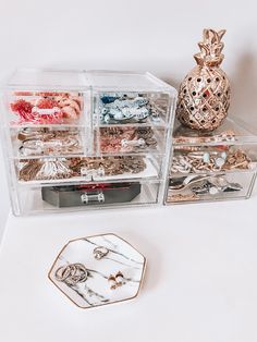 two clear boxes filled with jewelry sitting on top of a white table next to a gold pineapple