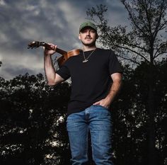 a man standing in front of trees holding a guitar and wearing a hat with his hands on his hips