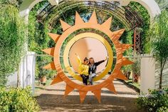 two people standing in front of an orange and yellow sun sculpture at the entrance to a garden