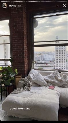 an unmade bed sitting in the corner of a room with large windows overlooking buildings