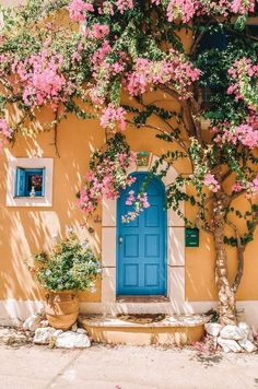 a blue door and some pink flowers on a yellow building with trees in the foreground