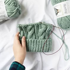 someone is knitting yarn on the bed with their green hand knitted hat and two balls of yarn