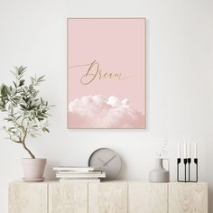 a pink and gold print with the word dream above it on a white wall next to a potted plant