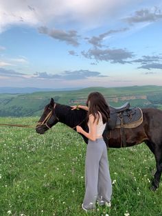 a woman standing next to a black horse on top of a lush green field under a cloudy sky