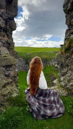 a woman sitting on the ground in front of an old stone wall with grass and rocks