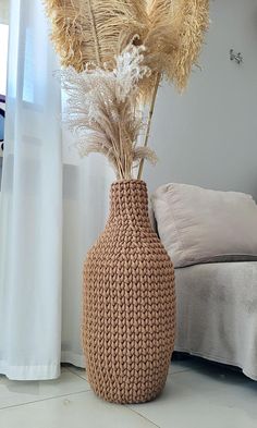 a crocheted vase with dried grass in it sitting on the floor next to a bed