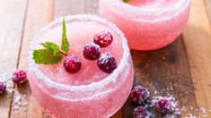 two glasses filled with ice and raspberries on top of a wooden table next to berries