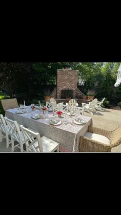 an outdoor dining table with white chairs and a fire place in the background, surrounded by greenery