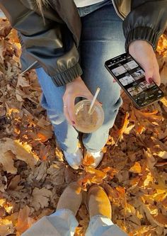 a person holding a drink and a cell phone in their hands while sitting on leaves