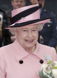 an older woman wearing a pink coat and black hat with flowers in her hand while standing next to other people