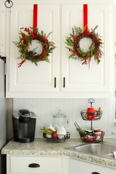 two christmas wreaths hanging on the wall above a kitchen sink with red ribbon around them