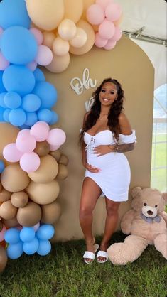 a woman in a white dress standing next to balloons and a teddy bear on the grass