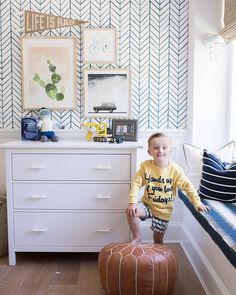 a young boy sitting on top of a brown ottoman in front of a white dresser