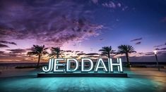 a neon sign that reads jedddah in front of palm trees and the ocean