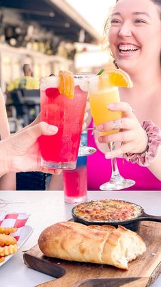 two women are toasting with drinks and pizzas on the table in front of them