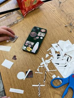 someone is cutting out paper with scissors and glue on a wooden table next to a cell phone