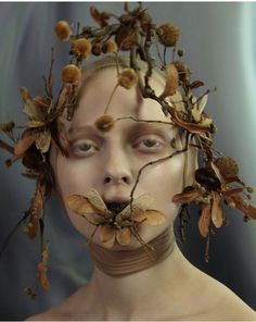 a woman with leaves on her head and branches attached to her face, in front of a blue background