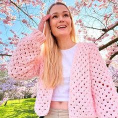 a woman wearing a pink crochet cardigan standing in front of a tree