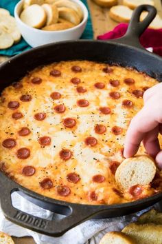 a hand dipping a piece of bread into a skillet filled with cheese and pepperoni