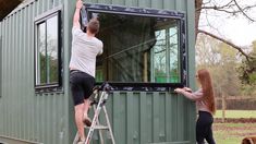 a man and woman are painting the windows of a green shipping container