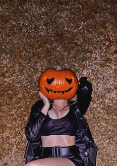 a woman wearing a black leather outfit and holding a pumpkin over her head while laying on the ground