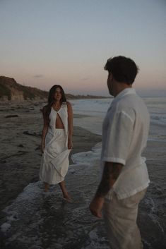 a man and woman walking on the beach at sunset, with one wearing a white dress