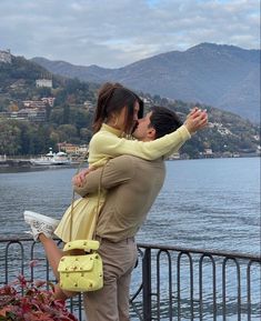 a man and woman are kissing by the water with mountains in the backgroud