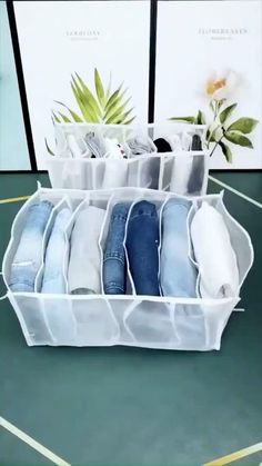 several pairs of jeans are stacked in a storage bin on the floor next to a plant