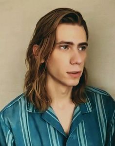 a young man with long hair wearing a blue shirt and standing in front of a wall