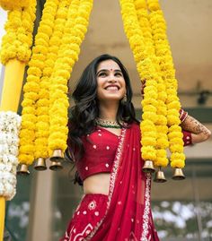 a woman in a red sari is standing under yellow flower garlands and smiling