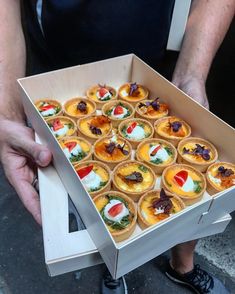 a man is holding a box full of small pastries with toppings on them