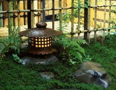 a lantern is sitting in the grass near some rocks and plants, with a bamboo fence behind it