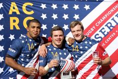 three men standing next to each other in front of an american flag banner holding drinks