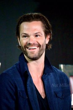 a man with a beard smiles at the camera while standing in front of a dark background