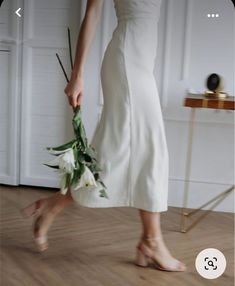 a woman in a white dress holding a bouquet of flowers and walking across a hard wood floor