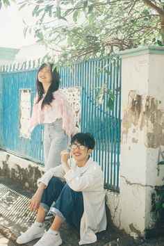 two people sitting on the ground next to a blue fence and some trees in front of them