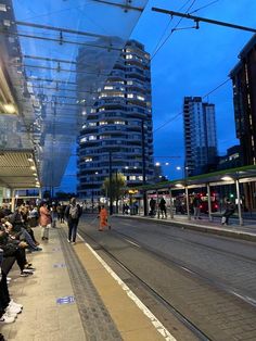 many people are sitting on the sidewalk in front of some tall buildings at night time