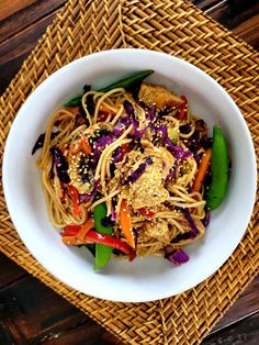 a white bowl filled with noodles and vegetables on top of a wooden table next to a woven place mat