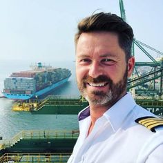 a man in uniform standing next to a large cargo ship on the water and smiling at the camera