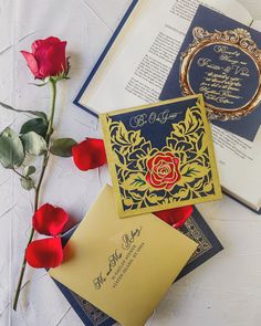 an open book and some red roses on a white table with blue and gold wedding cards