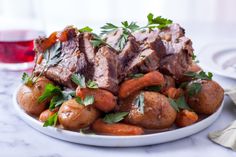 a white bowl filled with meat and potatoes on top of a marble table next to a glass of red wine