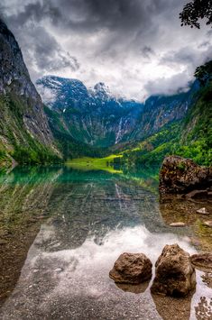 a lake surrounded by mountains and trees under a cloudy sky with rocks in the foreground