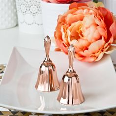 two small copper bells on a white plate next to a pink flower and potted plant